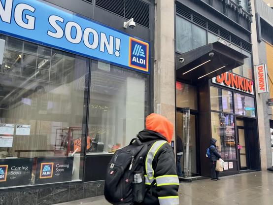 NYC sidewalk. Man crossing in foreground. Seen is a coming soon, Aldi’s, sign, beside an existing Dunkin’.