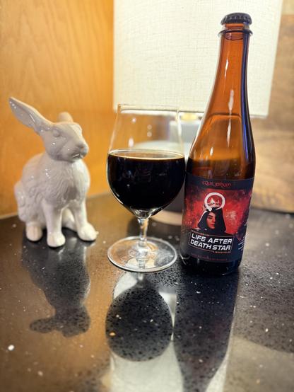 A white rabbit looks on quizzically as a snifter glass of dark beer and a pint bottle sitting front of a bright light on a shiny black countertop.