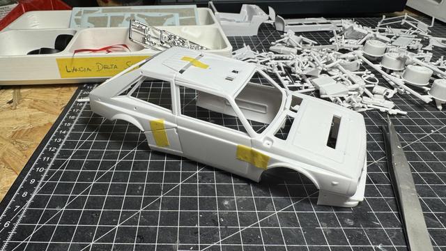 Nunu Beemax Lancia Delta S4 model kit body shell assembly in white with yellow masking tape, positioned on a cutting mat surrounded by unassembled white plastic sprues and parts containers