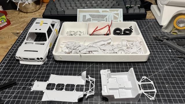 Nunu Beemax Lancia Delta S4 model kit contents organized in white compartmented trays, showing body shell assembly on the left, separated parts including decals, red wiring, black tires, and detailed components in individual sections