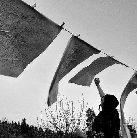 Image: photo, black-and-white; it appears to be of an artist drawing onto pieces of paper or fabric that is clothes-pegged to a clothes-line; they are blowing in the wind, and the artist is stretching up their right arm to draw; we only see from waist up of the artist; conifers and a bare tree or two in the background • “Curated by Eamonn Maxwell, Nósanna explores the transformative power of the ritual in Irish art and life. Eileen’s installations reimagine relics and Catholic rites from twentieth-century Ireland by assembling found objects and natural debris into delicate, hybrid artefacts. Arranged in curiosity cabinets, her forms commemorate and subvert traditional rituals, forging new meanings from discarded material. Gary’s “Wind Letters” channels the invisible language of the wind through meditative performances at sacred sites across the Midlands, using asemic writing and photography to capture fleeting presences tied to place and belief. His work, deeply influenced by the…”