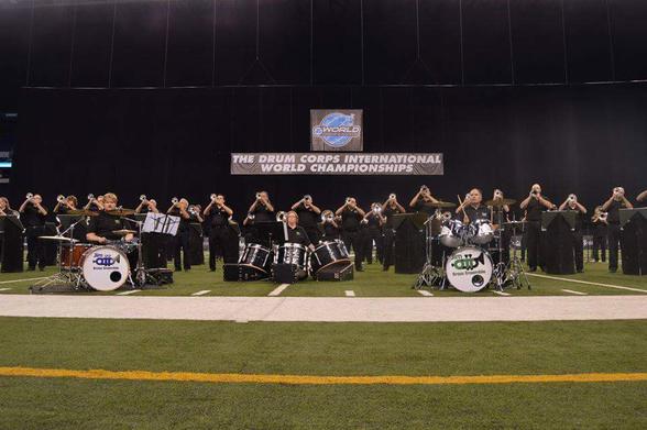The Jim Ott Brass Ensemble performing during the 2016 Drum Corps International World Championships at Lucas Oil Stadium in Indianapolis, Indiana. Two drum sets and a set of tuned marching bass drums are in front, while the rest of the the group is behind them in black attire, playing brass instruments.