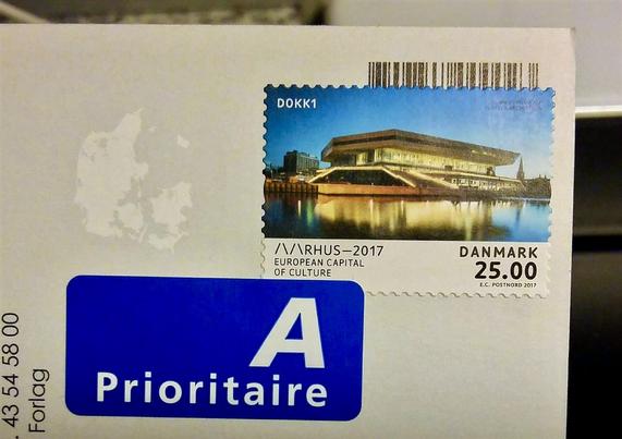 Close-up of the corner of an unposted postcard with an A-post air-mail sticker and a 25kr. stamp on it. The stamp features a photo of the waterfront Dokk1 Aarhus Main Library seen at night under a Blue Hour twilight sky with welcoming warm yellow light glowing from it. Underneath, the logo and text for Aarhus 2017 European Capital of Culture, which the stamp commemorates.