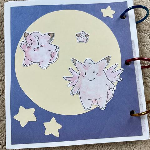 A photo of a page in my Pokédex scrapbook, featuring Clefairy and its evolution family. The background is made with cut out colored paper to look like a big full moon and stars