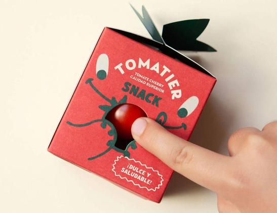 A hand presses a red snack box labeled "Tomatier Snack," featuring a playful design with eyes and a tomato-shaped opening.