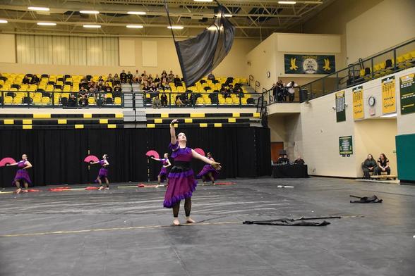 A color guard performance in a gymnasium featuring performers in purple costumes. One performer has tossed a large black flag, while others in the background are using red fans. Spectators are seated in the bleachers.