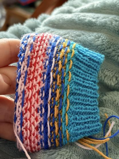 The inside of a very colorful stranded colorwork sock in progress. The cuff is a bright turquoise, the colorwork combines a mustard, light pink, cobalt blue and red.