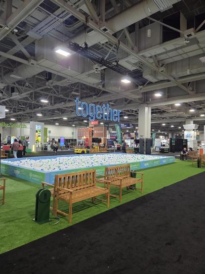 A large indoor convention hall is shown, with high industrial ceilings full of metal beams, air ducts, and bright overhead lights. Suspended above the center of the space is a glowing blue neon sign that reads **“together.”**
Below the sign is a large rectangular area filled with hundreds of small white plastic balls, with scattered blue and green ones mixed in. The ball pit is bordered by low panels decorated with the words **“RootsTech by FamilySearch”** and colorful graphics. The area around it is covered with bright green artificial turf, giving it the look of a small park inside the convention center.
In the foreground, two simple wooden benches sit on the turf facing the ball pit, as if inviting visitors to sit and watch or rest. A small dark-green post or stand is near the benches.
Beyond the ball pit are multiple exhibit booths and displays related to genealogy and family history. Large signs read **“FamilySearch,” “Storied,”** and **“Find your past.”** A few people are scattered around the space—some sitting on benches, others walking or talking at booths.
The overall feeling is open and collaborative, like a family-history expo or technology conference designed to bring people together to explore ancestry and storytelling.