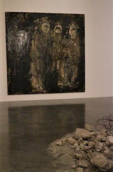 One large painting, apparently grey, textured figures on a white wall, on the floor a reflection of the painting, & some dust and chunks of concrete.