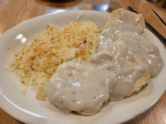 A white plate on a wooden table holds a Southern-style breakfast. Two fluffy biscuits are covered in thick white sausage gravy speckled with black pepper. Beside them is a pile of crispy golden hash browns. A glass of water sits blurred in the background, suggesting a cozy diner setting.