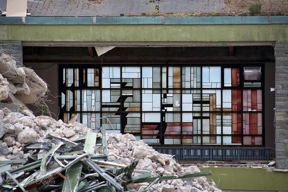 This image shows a partially destroyed building. In the foreground, there is a large pile of rubble consisting of broken bricks, concrete chunks, and twisted metal rods. The debris suggests significant structural damage.

Behind the rubble, you can see the remains of a building's facade. The structure has large, multi-paned windows, many of which are shattered or missing. The interior appears to be gutted, with visible damage to the floors and walls inside. The upper part of the building still has some intact sections, including a green-painted edge along the rooftop, but it looks unstable and heavily affected by whatever caused the destruction.

The scene evokes a sense of abandonment and damage, possibly from a natural disaster or demolition.