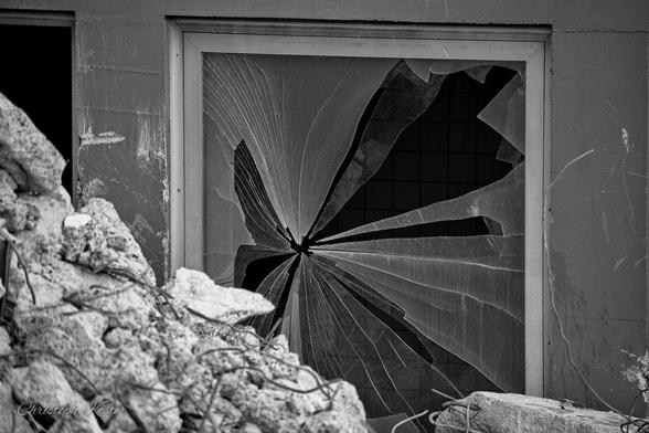 This black-and-white photograph captures a striking scene of destruction and decay. The focus is on a large, shattered window frame in the center of the image. The glass is broken, with sharp shards radiating outward from a central impact point, creating a dramatic, almost explosive pattern.

The window is part of a deteriorating building, with visible cracks and damage to the surrounding walls. To the left of the window, a section of the wall appears to have collapsed, revealing debris and rubble on the ground. The overall scene conveys a sense of abandonment and the aftermath of significant damage, possibly from an explosion or severe structural failure.
