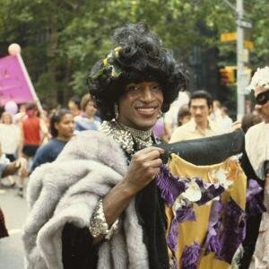 Marsha P. Johnson (1945-1992) pictured here in an undated color photograph at a Pride Event. She is a tall Black woman with a giant curly wig, diamond choker necklaces, a black cocktail dress, "mink" stole and diamond bracelets. She is smiling, and carrying part of a banner which cannot be seen. There are crowds on the sidewalk in the background along a tree lined road, and people marching behind her in the street.