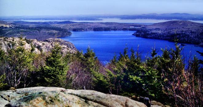 Beneath a clear blue sky we are standing on a light gray granite outcrop atop a mountain, looking out over the tops of a mixed forest of evergreens and leafless hardwoods running down the mountainside before us. Just beyond the trees on our left and just below us is a rocky crag with only a few stunted pines growing from crevices along its crest. A large deep blue pond dominates the view below in front of us. To our right a small rounded mountain rises from the pond's shore. Beyond the pond is a narrow, flat wooded strip of land backed by a long ocean inlet and then another relatively fat piece of land  with a range of mountains to our right and scattered mountains along the horizon.