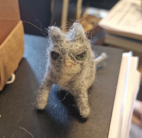 A grey cartoonish cat made of felt. He looks kinda fat with skinny legs and grumpy