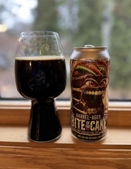 A stout glass full of dark beer sits on a window sill next to a gold pint can with a renegade piece of cake, teeth bared, spicy chilis in its hair.