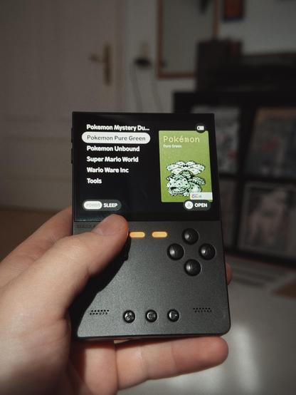 A person holding a portable gaming device displaying a selection menu for Pokémon games, with a particular focus on Pokémon Pure Green.