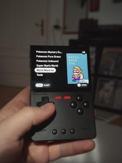 A handheld gaming device displaying a list of game titles and a pixel art image of Wario on the screen.