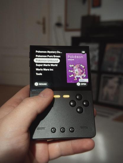 Hand holding a portable gaming device displaying a game selection menu with "Pokémon Unbound" highlighted.