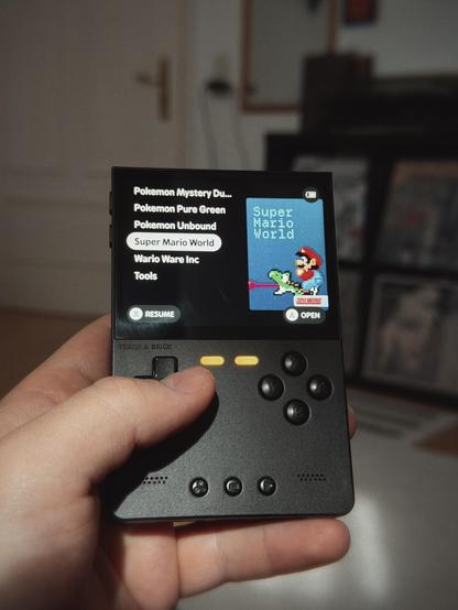 A handheld gaming device displaying a list of game titles, with "Super Mario World" selected and a pixelated Mario image on the screen.