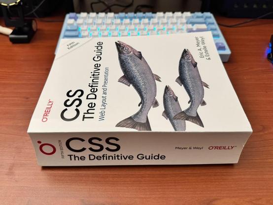 Photo of the ginormous 5th Edition of CSS: The Definitive Guide by Eric A Meyer and Estelle Weyl published by O'Reilly Books. It's sitting on a desk in front of a powder blue mechanical keyboard.