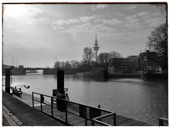 Black-and-white waterfront scene along the River Geeste. In the foreground is a small dock with railings, beyond it calm water. A bridge spans the river on the left, buildings line the right bank, and a tall tower rises in the distance under a lightly clouded sky.