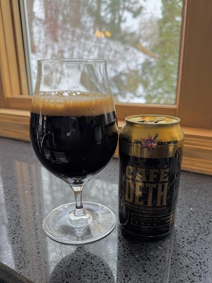 A large snifter glass full of dark beer with a tan head sits on a window sill with a shiny black and gold can. Cafe Deth is spelled out in gold letters. A snowy scene is in the background