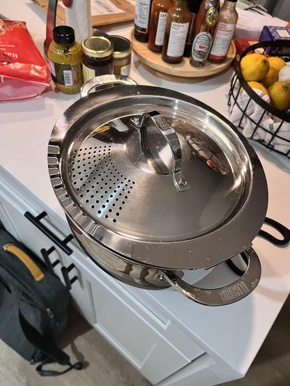 A close-up, high-angle shot depicts a shiny, stainless steel pot and lid, featuring a perforated strainer section on the lid. The lid has a large handle branded with the word "BIALETTI", and the pot is set on a white surface, with other kitchen items slightly visible in the background, including a wire basket of lemons, a tray of sauce bottles, and jars.