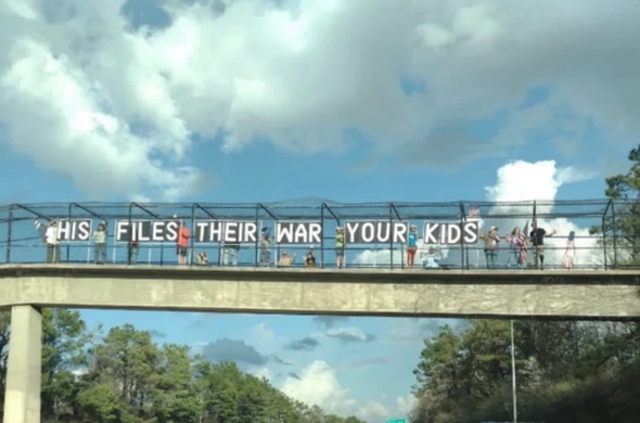 Picture from a car looking up at a fenced-in overpass. Protesters have put up all-cap letters reading "HIS FILES THEIR WAR YOUR KIDS".