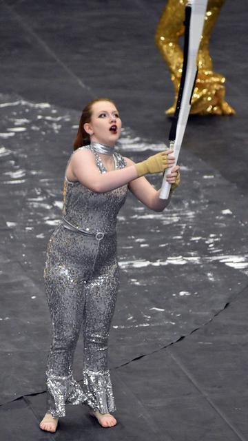 A performer in a shiny silver costume stands on a stage, holding a white rifle. The individual appears to be engaged in a dynamic performance, with an expression of enthusiasm. In the background, another performer in a gold costume is partially visible.