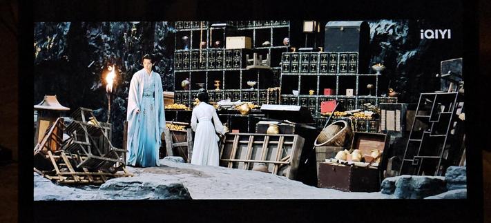 Hong Ye and Xiao Yao standing in a large treasure chamber with lots of furniture strewn about - apparently support for a lot of gold. The decorated shelving unit sports a geomeyrical gold pattern on black.