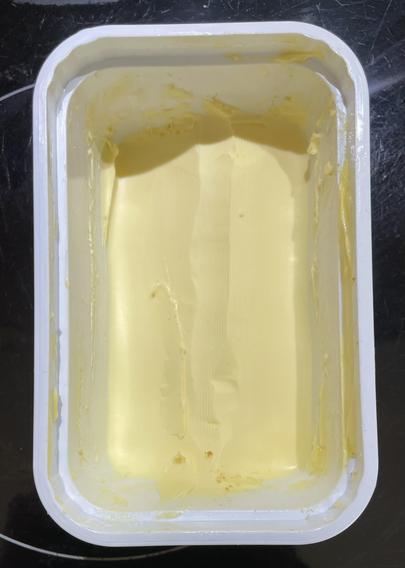 Overhead shot of an open tub of yellow spreadable butter (butter and margarine blend), with a pretty regular pattern of use…