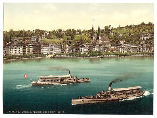 The image depicts a picturesque scene of the Swiss cityscape with historical charm. In the foreground, two steamboats are seen cruising on calm blue waters. The boat closer to the viewer is marked "Promenade & HOFKIRCHE" and bears a prominent red flag bearing a white cross, indicating its association with Switzerland's national symbol.
The backdrop showcases an array of European-style buildings lining the waterfront in Lucerne. They feature traditional architecture reminiscent of the region’s history, including ornate facades and steep roofs characteristic of Alpine towns. Further back, atop rolling green hills, lie two tall spires that appear to be part of a prominent church or cathedral.
The overall impression is one of leisurely travel through an idyllic European setting during the late 19th century, with historical vessels adding character to the scene.