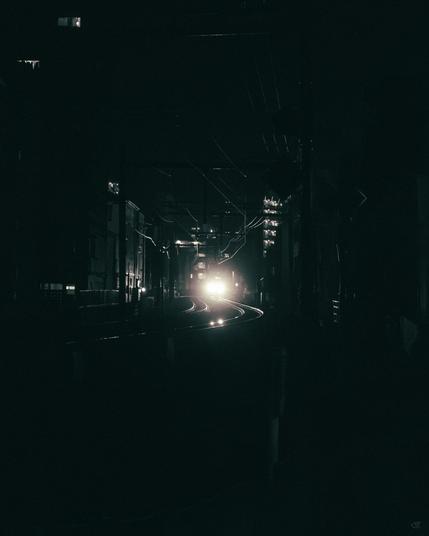 Dark nighttime view down a narrow urban rail corridor, with curving train tracks leading toward bright headlights in the distance, buildings and utility wires lining both sides, and light rain streaks visible.