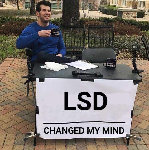 Meme image with text. The photo is a guy sitting at a table, holding a coffee. On the table are some papers and peripherals and in front hangs a banner with the text: LSD - changed my mind.