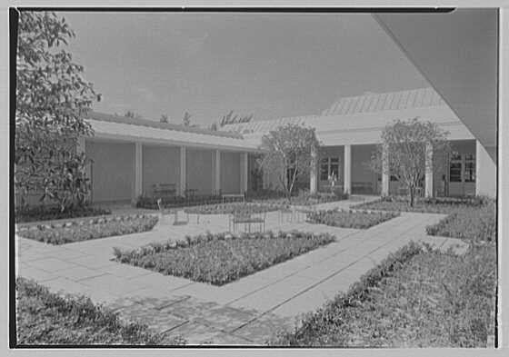 This black and white photograph depicts a courtyard area of what appears to be an art gallery or educational institution. The image shows a series of columns supporting the structure's roof, creating covered areas along the sides of the building. In between these columned sections are open spaces with neatly arranged benches for sitting.
The central focus is on a large tiled patio surrounded by manicured lawns and flower beds that add to the overall aesthetic appeal of the space. The pathways leading into this area are paved in stone, directing visitors towards different parts of the facility or gardens around it.
In summary, the image captures an organized and visually pleasing outdoor setting at what seems like a cultural institution built during 1941 March.