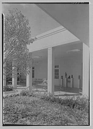 This black and white photograph shows a large, open structure with pillars supporting the roof. The building appears to be an exhibition or gallery space as there are several sculptures displayed on pedestals within it. Outside of the structure, in what looks like a courtyard area, is sparse grass and some trees providing partial shade. It seems to capture a moment at the Norton Gallery & School of Art located in West Palm Beach, Florida during March 1941 according to archival records from Gottscho-Schleisner Inc., suggesting this might be part of their collection or project documentation.