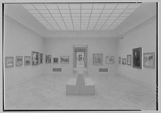 The image displays a spacious gallery with an expansive view of art on the walls. The room is well-lit, presumably by artificial lighting given its uniform illumination across all areas without any discernible windows or natural light sources.
Numerous framed artworks are neatly arranged in rows along both sides of the central pathway that runs through the middle of the space. Each artwork appears to be hung at eye level for comfortable viewing, and there is ample spacing between them, allowing visitors easy access to each piece.
In the foreground, a large bench sits parallel to one wall on either side, providing seating options with its elongated form accommodating multiple people comfortably. The flooring consists of broad wooden planks that add warmth to the otherwise neutral-toned gallery walls.
At the far end, beyond what seems like an open door or entrance, more artwork can be glimpsed in a slightly darker space, suggesting additional rooms for viewing within this gallery setting. Overall, the image conveys a sense of calm and contemplation typical of art exhibitions designed with visitor experience at heart.