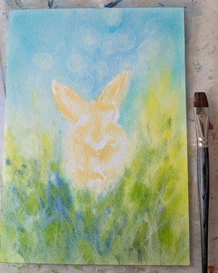 Work in porgress shots of my new bunny portrait - A cute rabbit with brown fur sits in a green meadow under a blue sky. It looks curiously out from among yellow wildflowers towards the viewer. Here an early stage of the painting with the first layers of watercolor paint.