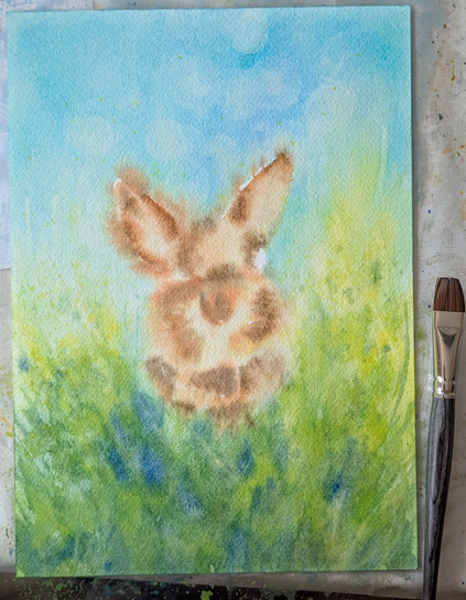 Work in porgress shots of my new bunny portrait - A cute rabbit with brown fur sits in a green meadow under a blue sky. It looks curiously out from among yellow wildflowers towards the viewer. Here another stage of the painting with more layers of watercolor paint.