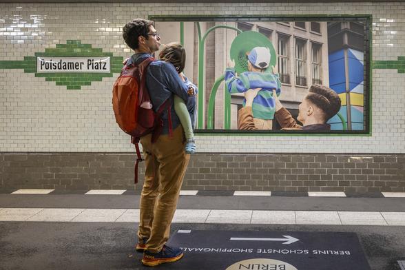 A street photography scene on a metro platform in Berlin: A dad is holding his young daughter in his arms while waiting for the next train. She is sleeping and has her head burried in his shoulder, one leg covered in a green legging hanging on one side. On the wall opposite, a frame with a big photograph of one dad holding his son up in his arms so that he can speak into a green megaphone. The little boy also wears a green and blue striped sweater. An old enamel sign next to the picture reads "Potsdamer Platz".