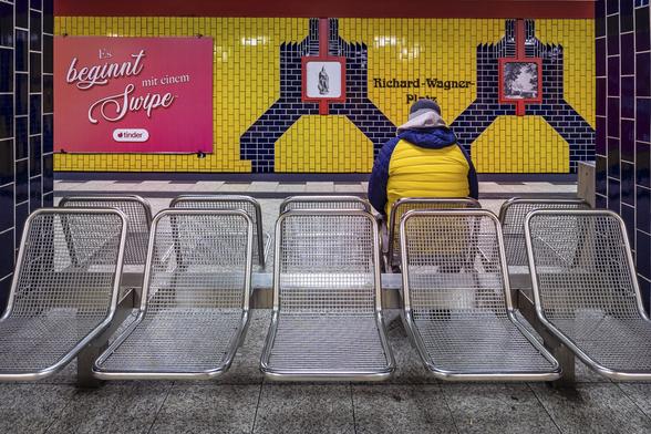 A street photography scene on a metro platform in Berlin: A man with a bright yellow down jacket with dark blue arms is sitting on a chair facing a wall covered in tiles of the same bright yellow colors and with geometric shapes in dark blue tiles. The man is almost invisible against the wall like a cameleon as his arms fit perfectly between two 'legs' of the shape. Above his head, in dark tiles, the words "Richard Wagner Platz". On his left on the wall, a commercial poster for the Tinder website.