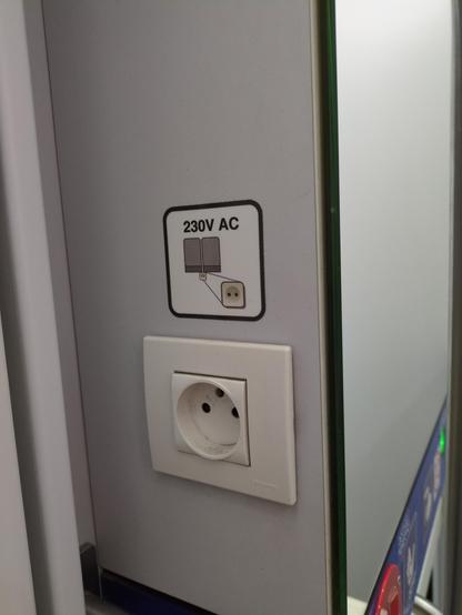 Gniazdko elektryczne na boku pudła lustra w łazience pociągu. Nad nim etykieta z piktogramem. U góry tekst "230V AC". Poniżej rysunek przedstawiający dwa przylegające do siebie szare prostokąty, lekko zaokrąglone u góry, z ciemniejszymi pasami na dole. Poniżej na środku mała, grafika gniazdka w kolorze kremowym, która następnie jest powiększona ze wskazaniem oryginalnej pozycji.