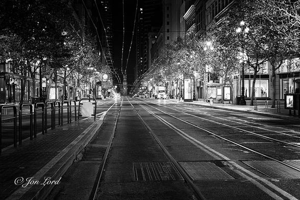 This is a black and white nighttime photo in landscape format looking along a pair of city tram tracks. San Francisco, California (2012).
The camera is positioned about 1m above the street and looking along along a straight stretch of tram tracks. The tracks are imbedded in the tarmac of the road. To the right is a second set of parallel rails. Suspended above the road are the electric cables that power the trams, these are shiny and reflect the light from the street below. The pavement on either side of the road is lined with mature trees that are covered in Christmas lights. Behind the pavement are the illuminated shop fronts of a city centre. The street is deserted.