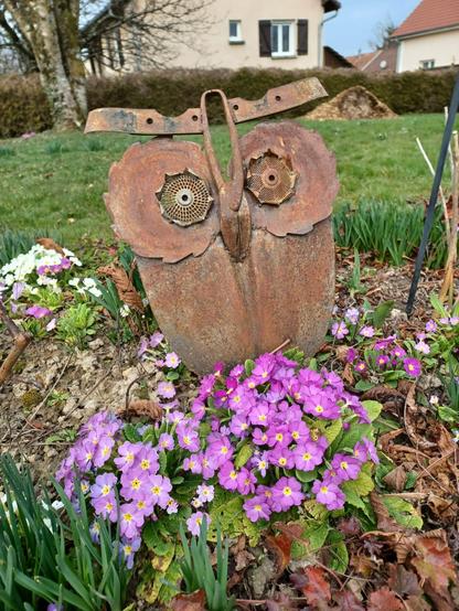 Chouette réalisée à partir d'une pelle, 2 lames de scies circulaires et 2 grilles de moulin à légumes pour les yeux, morceaux d'outils agricoles pour les sourcils, une grosse pince coupante pour le bec, posée dans un jardin au milieu des primevères violettes