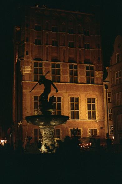 Color photo of Neptunes fountain at old town in Gdansk, PL. The photo has been taken at night, the fountain statue is in shadows, the building behind is illuminated