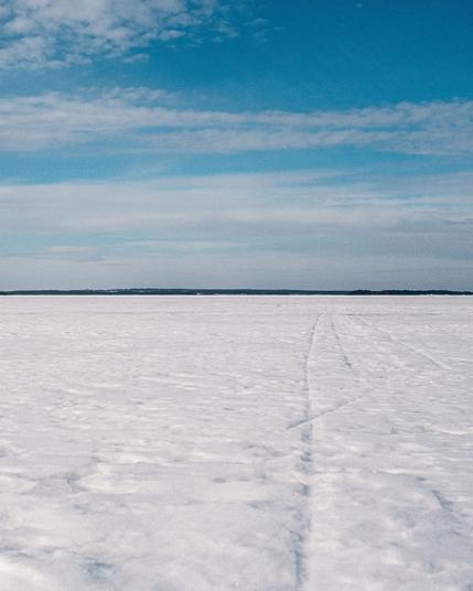A vast frozen lake stretches to the horizon under a bright blue sky scattered with thin white clouds. Two parallel tracks — likely from a snowmobile or skis — cut through the snow-covered ice, converging toward a distant dark treeline.