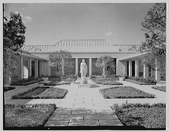 The image depicts a symmetrical and formal garden setting, with neat rows of trimmed hedges on either side. In the center is a statue standing upright on what appears to be an elevated pedestal or platform within a paved walkway leading up to it. The building in front features classical architectural elements such as columns supporting a roof structure that covers most of its length and has large windows, likely providing ample natural light for indoor activities or display purposes. Trees are visible at the edges of the courtyard, framing this peaceful outdoor environment.
The photograph is monochromatic, suggesting an older timeframe when black-and-white film was commonly used in photography. The absence of color adds a sense of timelessness and perhaps reflects upon the historical context from which it originated: January 17, 1942.