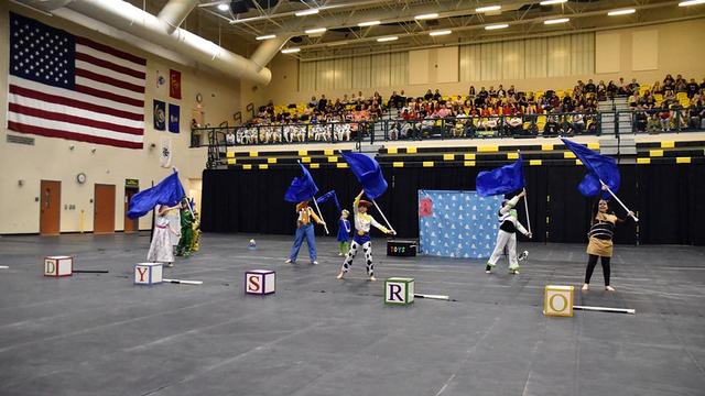 A performance scene featuring individuals dressed in Toy Story costumes, waving blue flags. They stand among large, colorful letter blocks on the floor. In the background, an audience watches from bleachers, with an American flag displayed on the wall.