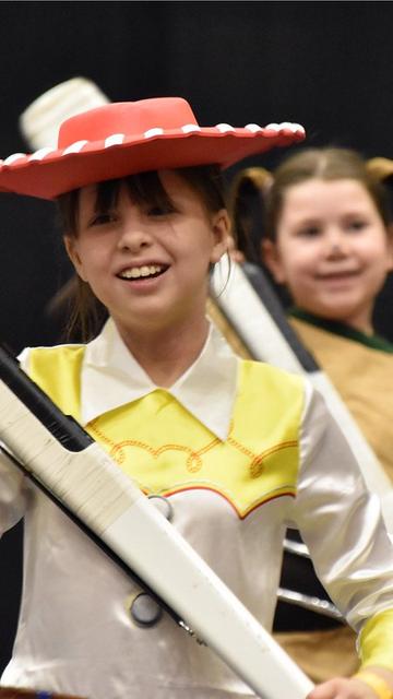 Two children are performing in costumes; one girl is dressed as Jessie from Toy Story, smiling while holding a wooden rifle. The background is dimly lit, focusing on their joyful expressions.